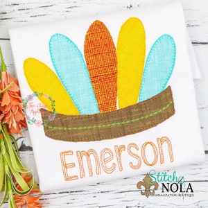 May include: White t-shirt featuring an embroidered Native American headdress design. The headdress has yellow, blue, and orange feathers, with the name "Emerson" embroidered in orange. The design includes the text "Stitch NOLA Personalization Boutique".