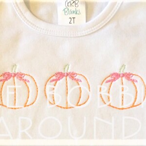 May include: White toddler shirt with three embroidered orange pumpkins with pink bows. The text "Three Pumpkins Around" is embroidered in white below the pumpkins.