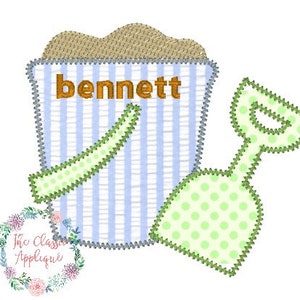 May include: A blue and white striped bucket with brown sand and the name "bennett" on the side. A green and white polka dot shovel is next to the bucket.