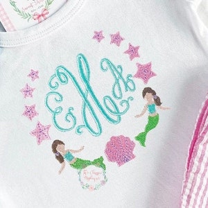 Puede incluir: Un pelele blanco con un monograma bordado turquesa y un diseño de dos sirenas y estrellas de mar. El pelele se combina con pantalones cortos de rayas rosa y blancas.