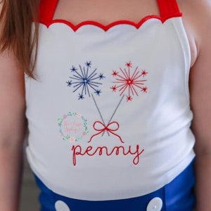 K&ouml;nnte beinhalten: Wei&szlig;es &auml;rmelloses Shirt mit rotem Besatz und einem gewellten Ausschnitt. Das Shirt zeigt ein gesticktes Design aus zwei roten und blauen Feuerwerksk&ouml;rpern mit einer roten Schleife und dem Namen "penny" darunter.