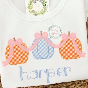 May include: A white t-shirt with three embroidered pumpkins in a row. The pumpkins are orange and white gingham with pink bows. The word "harper" is embroidered in blue below the pumpkins.