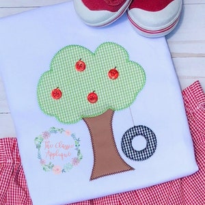 May include: A white onesie with a green and brown applique of an apple tree with a tire swing. The onesie is paired with red and white gingham shorts.