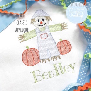 May include: A white fabric applique of a scarecrow with a straw hat, holding two orange pumpkins. The scarecrow is wearing blue overalls and has a smiling face. The text "The Classic Applique" and "Bentley" are embroidered below the scarecrow.