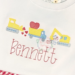 Op de afbeelding: Een wit T-shirt met een gele bouwtruck die een rood hart vervoert en een kleinere gele bulldozer met harten die eruit komen. De naam "Bennett" is geborduurd onder de trucks.