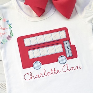 May include: White short-sleeved shirt with a red double-decker bus applique and the name "Charlotte Ann" embroidered in red thread.