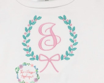 May include: A white baby onesie with a pink and teal embroidered wreath with a pink letter "S" in the center and a pink bow underneath. The text "The Classic Applique" is visible in the bottom left corner.