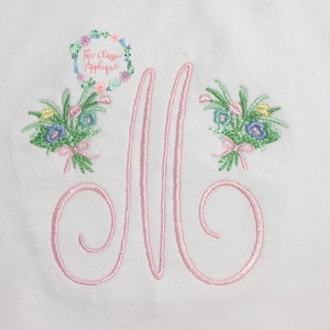 Op de afbeelding: Een geborduurd monogram met de letter "M" in roze, met aan weerszijden twee bloemboeketten. De boeketten zijn geborduurd met groene bladeren, roze, blauwe en gele bloemen.