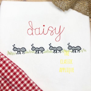 Op de afbeelding: Borduurwerk van de naam "daisy" in rode draad met vier zwarte mieren die over groen gras marcheren. De tekst "The Classic Applique" staat in gele draad onder de mieren.