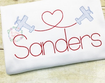 May include: A white embroidered shirt with a red heart and two blue airplanes flying in opposite directions. The text "Sanders" is embroidered in red below the airplanes.