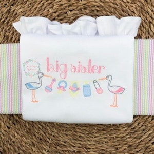 May include: White baby onesie with pink and blue embroidery. The onesie says "big sister" and features a stork carrying a baby rattle, pacifier, and other baby items.