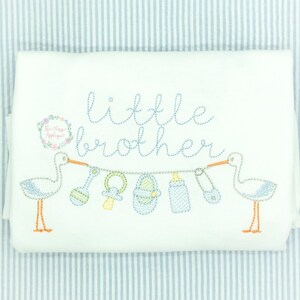 May include: White onesie with blue and white embroidered storks carrying a string of baby items, including a rattle, a pacifier, a shoe, a bottle, and a safety pin. The text "little brother" is embroidered above the storks.