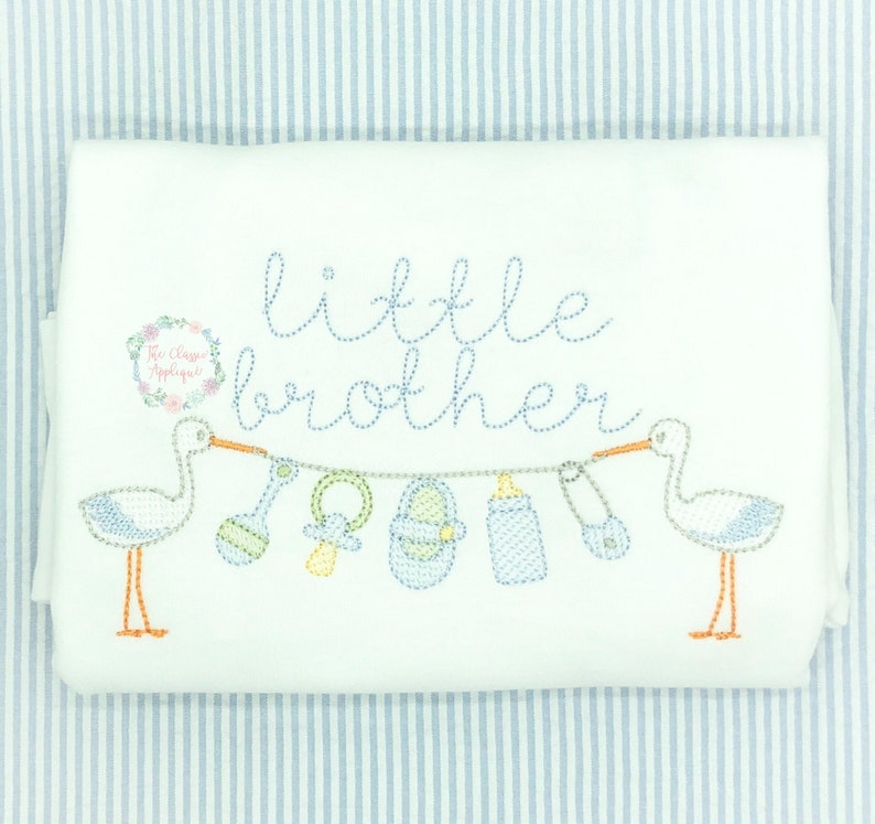 May include: White onesie with blue and white embroidered storks carrying a string of baby items, including a rattle, a pacifier, a shoe, a bottle, and a safety pin. The text "little brother" is embroidered above the storks.