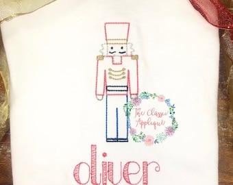 May include: A red and blue embroidered nutcracker with a gold trim and a wreath with the text "The Classic Applique" in pink. The name "oliver" is embroidered in pink below the nutcracker.