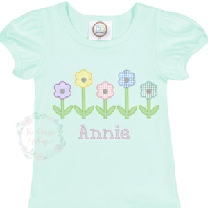 May include: A light blue t-shirt with a row of five embroidered flowers in different colors. The flowers are yellow, purple, pink, blue, and green. The name "Annie" is embroidered below the flowers.