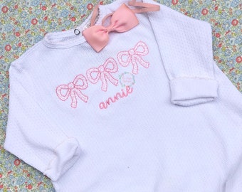 May include: White baby onesie with pink embroidered bows and the name "annie" on the front.