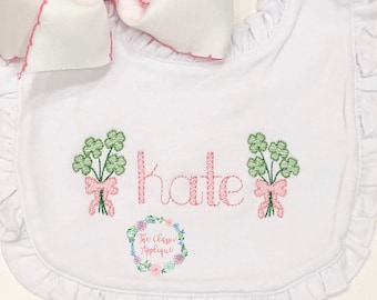 May include: White bib with pink ruffled trim and green shamrock embroidery. The name "Kate" is embroidered in pink. The bib has a small logo for "The Classic Applique" in the bottom center.