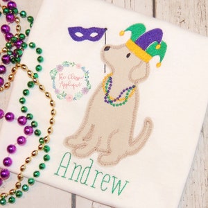 May include: A white t-shirt with an embroidered image of a dog wearing a purple and green jester hat, a purple mask, and a purple, green, and gold beaded necklace. The dog is holding a purple mask on a stick. The name "Andrew" is embroidered in green below the dog.