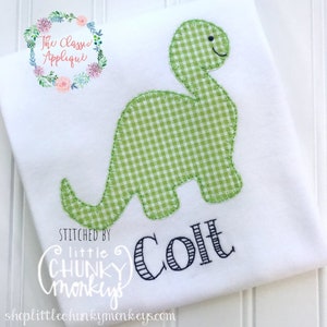 Puede incluir: Un pelele blanco con un aplique de dinosaurio de vichy verde y el nombre "Colt" bordado en hilo negro. El pelele está cosido por "little chunky monkeys".