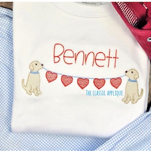 May include: White baby onesie with the name "Bennett" embroidered in red. Two yellow dogs are holding a string of red hearts.