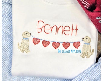 May include: White baby onesie with the name "Bennett" embroidered in red. Two yellow dogs are holding a string of red hearts.