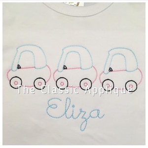 May include: A white t-shirt with three embroidered cars in a row. The cars are pink, blue, and blue. The text "The Classic Applique" and the name "Eliza" are embroidered on the shirt.
