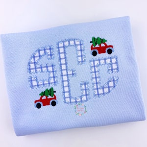 May include: A light blue baby onesie with a monogram of the letters "SEC" in a blue and white plaid pattern. The letters are outlined in blue stitching. Two red toy trucks with green Christmas trees are embroidered on the onesie.