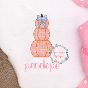 Puede incluir: Una pila de tres calabazas naranjas con un lazo azul en la parte superior. Las calabazas están bordadas en una tela blanca con el nombre "penelope" escrito en rosa debajo.