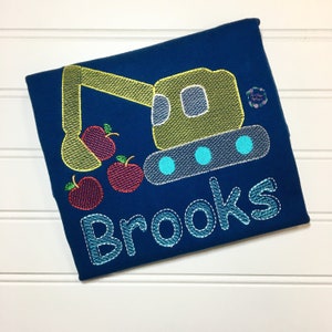 May include: A blue t-shirt with an embroidered design of a yellow excavator digging up red apples. The name "Brooks" is embroidered below the excavator.