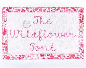 May include: A white fabric with a pink floral border and the text "The Wildflower Font" embroidered in pink thread.