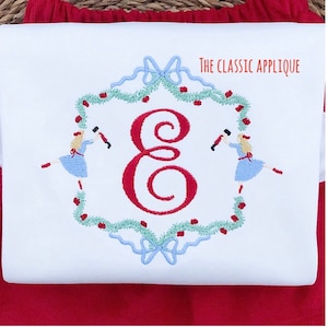 May include: White embroidered appliqué with a red letter E, two figures in blue dresses holding red and black nutcracker figures, and a green garland with red berries. The text "THE CLASSIC APPLIQUE" is in the top right corner.