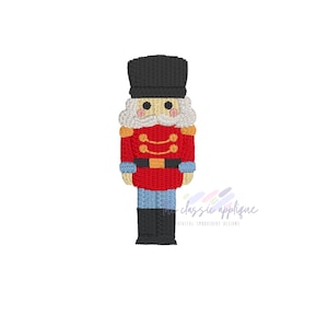 May include: A colorful embroidered design of a toy soldier nutcracker. The nutcracker is wearing a red jacket with gold accents, blue pants, and a black hat. The design includes the text "classic applique".