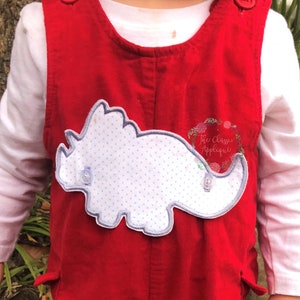 Peut inclure: Une salopette en velours côtelé rouge avec une applique de dinosaure bleue et blanche. Le dinosaure a un motif de pois blancs.