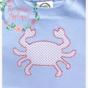 May include: A light blue t-shirt with a red embroidered crab applique. The crab has a white polka dot pattern.