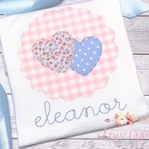 May include: A white t-shirt with a pink and white gingham heart design with two smaller hearts on top. The smaller hearts are floral and blue with white polka dots. The name "eleanor" is embroidered in blue thread below the hearts.