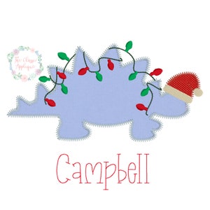 May include: A blue dinosaur with a red Santa hat and Christmas lights wrapped around it. The name "Campbell" is written below the dinosaur.