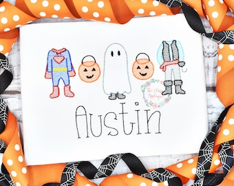 Op de afbeelding: Een wit T-shirt met een geborduurd ontwerp van drie kinderen gekleed in Halloween-kostuums: een superheld, een geest en een piraat. Elk kind houdt een pompoen-emmer vast. De naam "Austin" is onder de kostuums geborduurd.