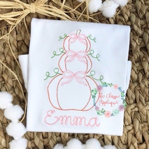 Puede incluir: Camiseta blanca bordada con una pila de tres calabazas con lazos rosas y enredaderas verdes. El nombre "Emma" está bordado debajo de las calabazas.