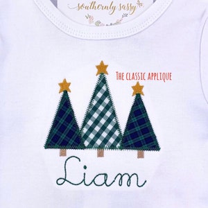 May include: White onesie with three Christmas tree appliqués. The trees are green and navy plaid and green and white gingham, topped with gold stars. The name "Liam" is embroidered in green script below the trees. Text reads "THE CLASSIC APPLIQUE".