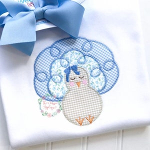 Puede incluir: Una camisa blanca con una aplicación azul y blanca de un pavo con un lazo azul. El pavo está sentado con los ojos cerrados. El texto "The Classic Applique" es visible en la esquina inferior izquierda de la imagen.