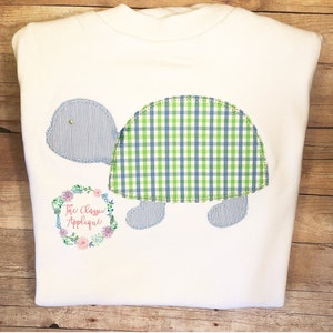 May include: White t-shirt with a blue and green checkered turtle applique. The turtle has a blue and white striped shell. The shirt also has a small floral wreath with the text "The Classic Applique" in pink.