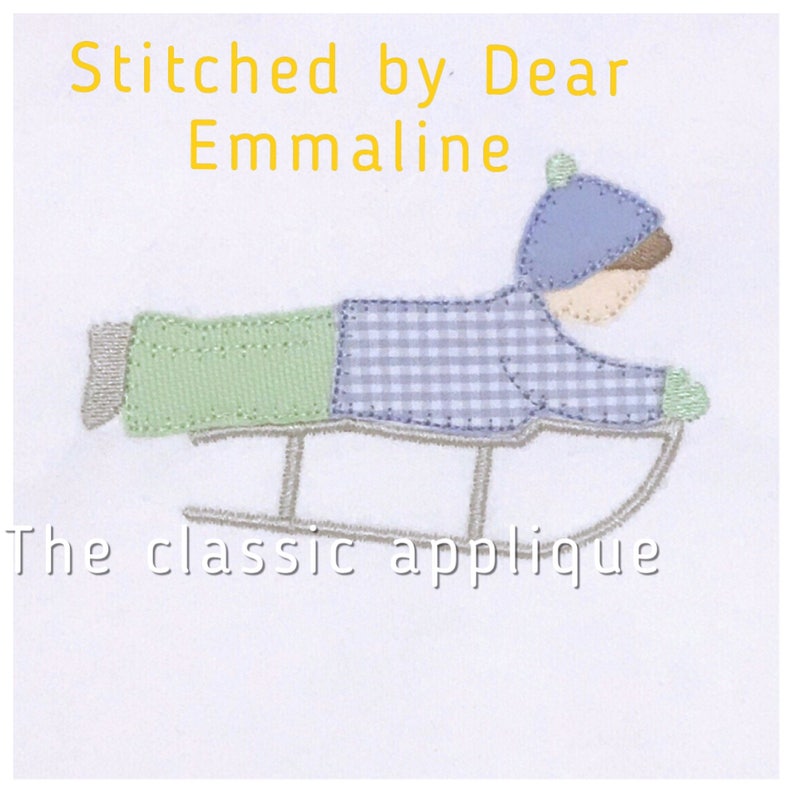 Pu&ograve; includere: Design di applicazione di un bambino che va in slitta, con cappello blu e camicia a quadri, pantaloni verdi e guanti. La slitta &egrave; grigia. Il testo recita "Stitched by Dear Emmaline" e "The classic applique".
