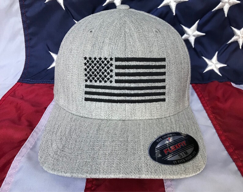 Op de afbeelding: Een grijze baseballpet met een zwarte geborduurde Amerikaanse vlag op de voorkant. De pet heeft een Flexfit-label op de achterkant, met de tekst "The Original Flexfit Sim It Fits Your Life".