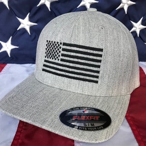 Op de afbeelding: Een grijze baseballpet met een zwarte geborduurde Amerikaanse vlag op de voorkant. De pet heeft een Flexfit-logo aan de zijkant en de tekst "The Original Flexfit SIM It Fits Your Life".