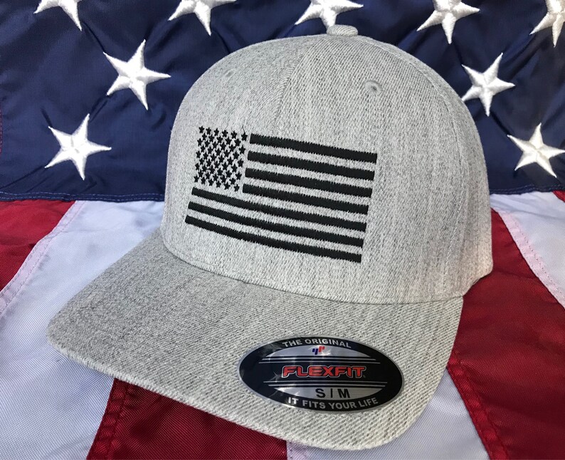 Op de afbeelding: Een grijze baseballpet met een zwarte geborduurde Amerikaanse vlag op de voorkant. De pet heeft een Flexfit-logo aan de zijkant en de tekst "The Original Flexfit SIM It Fits Your Life".