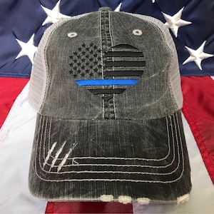 Puede incluir: Una gorra de camionero negra y gris desgastada con un dise&ntilde;o de bandera estadounidense en forma de coraz&oacute;n y una l&iacute;nea azul en el centro. La gorra est&aacute; hecha de un material de malla y tiene una parte posterior de malla blanca.