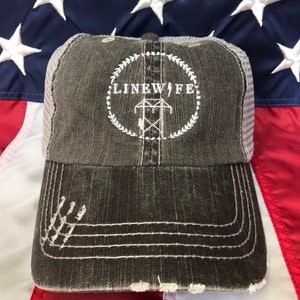Peut inclure: Un chapeau de camionneur gris et blanc délavé avec un logo brodé blanc qui dit "LINEWIFE" avec une tour de ligne électrique à l'intérieur d'un cercle de feuilles.