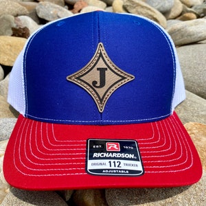 May include: A blue, white and red trucker hat with a brown leather patch featuring the letter "J" in a diamond shape. The hat is adjustable and has the text "EST. 1970 RICHARDSON. ORIGINAL 112 TRUCKER ADJUSTABLE" on the back.