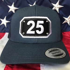May include: A dark blue trucker hat with a white mesh back. The hat features a black and white patch with the number "25" on it. The hat is made by Yupoong and is a snapback style.