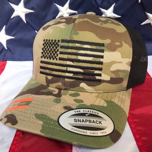 May include: A camouflage trucker hat with a black mesh back. The hat features an embroidered American flag with black stars and stripes. The hat also has a black patch with the text "THE CLASSICS SNAPBACK YUPOONG SINCE 1974".